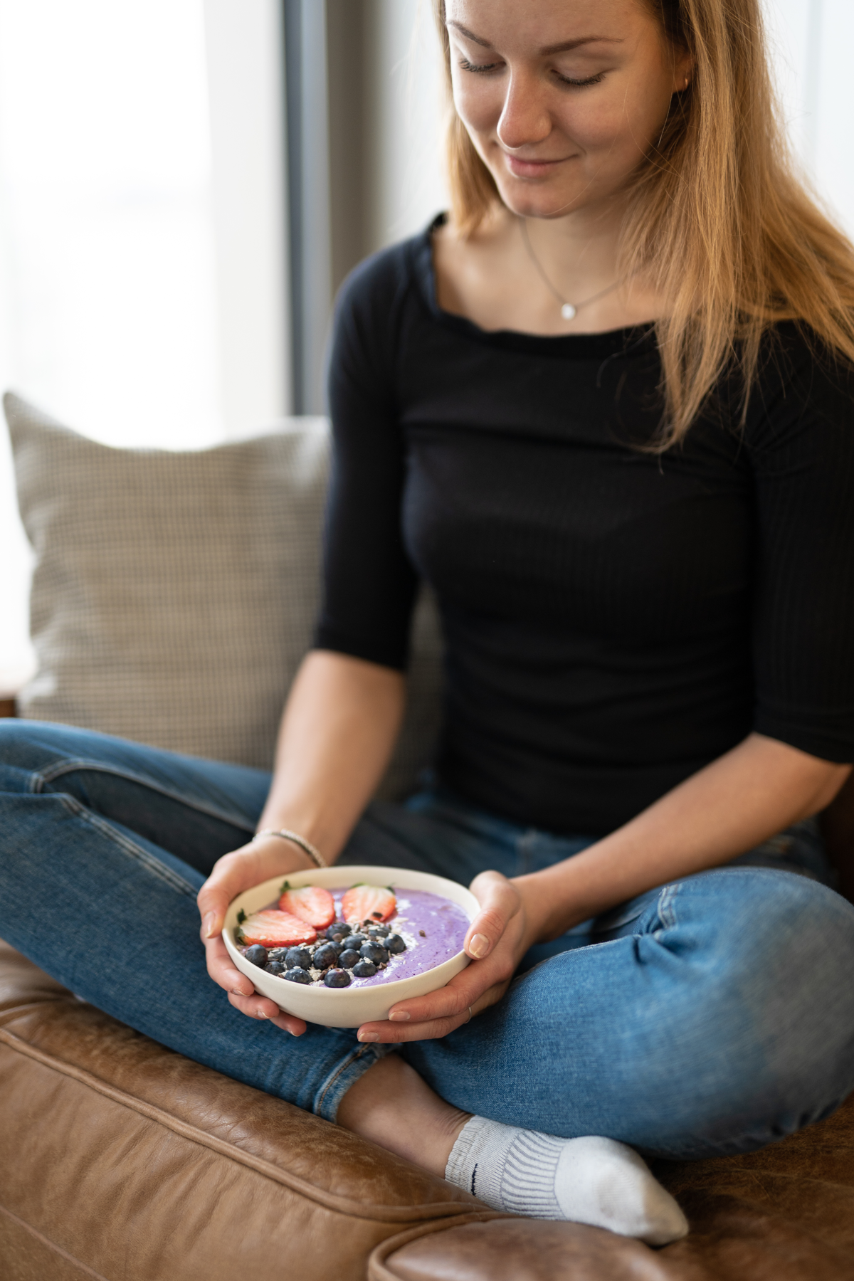 Person hält eine Schüssel mit violettem Protein-Smoothie-Bowl mit Erdbeeren und Blaubeeren, sitzt entspannt auf einem Sofa