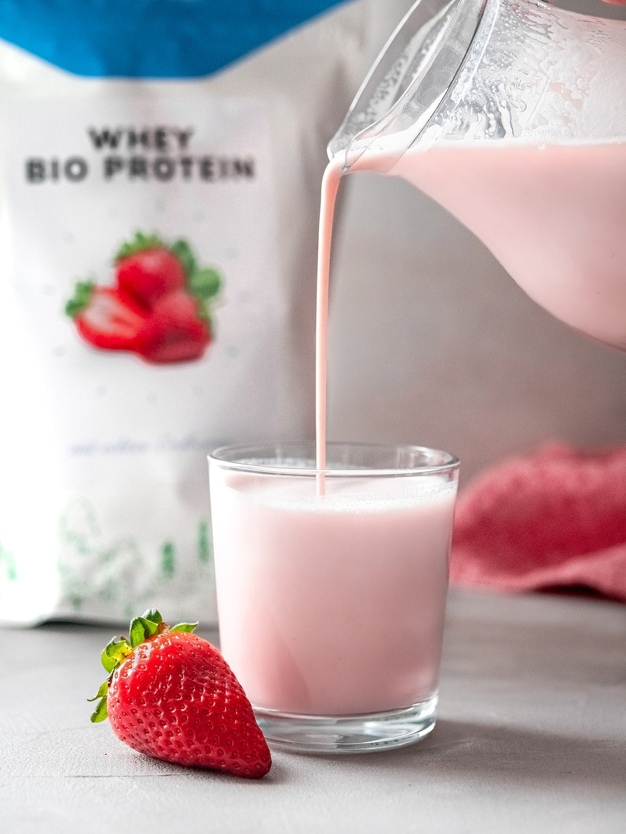 Erdbeer-Molkenprotein-Shake wird in ein Glas gegossen, mit frischer Erdbeere im Vordergrund und Sportbionier Whey Bio Protein Packung im Hintergrund.