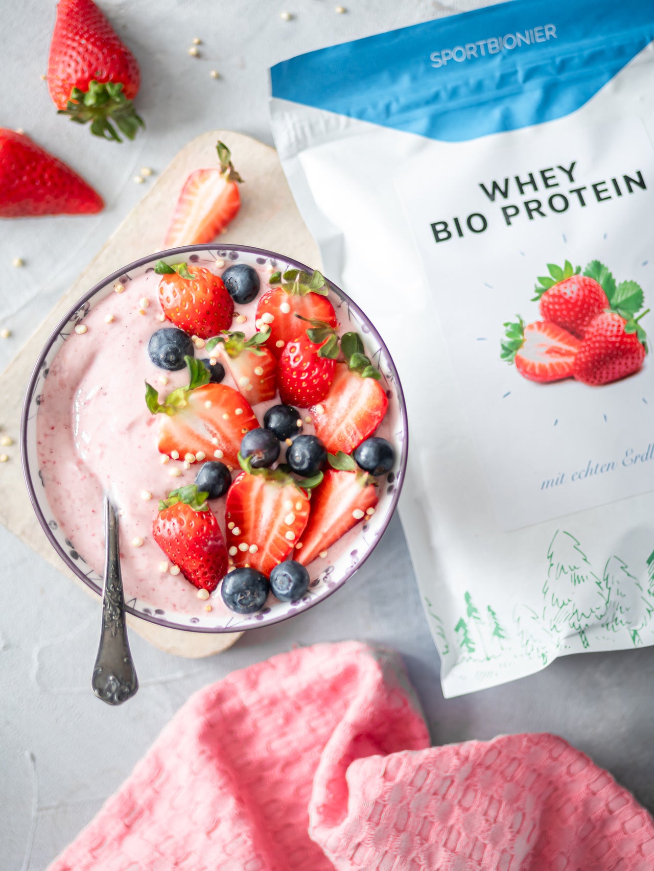 Schüssel mit cremigem Erdbeer-Protein-Smoothie-Bowl mit frischen Erdbeeren und Blaubeeren neben einer Packung Sportbionier Whey Bio Protein Erdbeere.