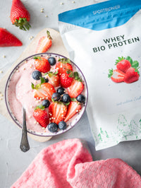 Schüssel mit cremigem Erdbeer-Protein-Smoothie-Bowl mit frischen Erdbeeren und Blaubeeren neben einer Packung Sportbionier Whey Bio Protein Erdbeere.